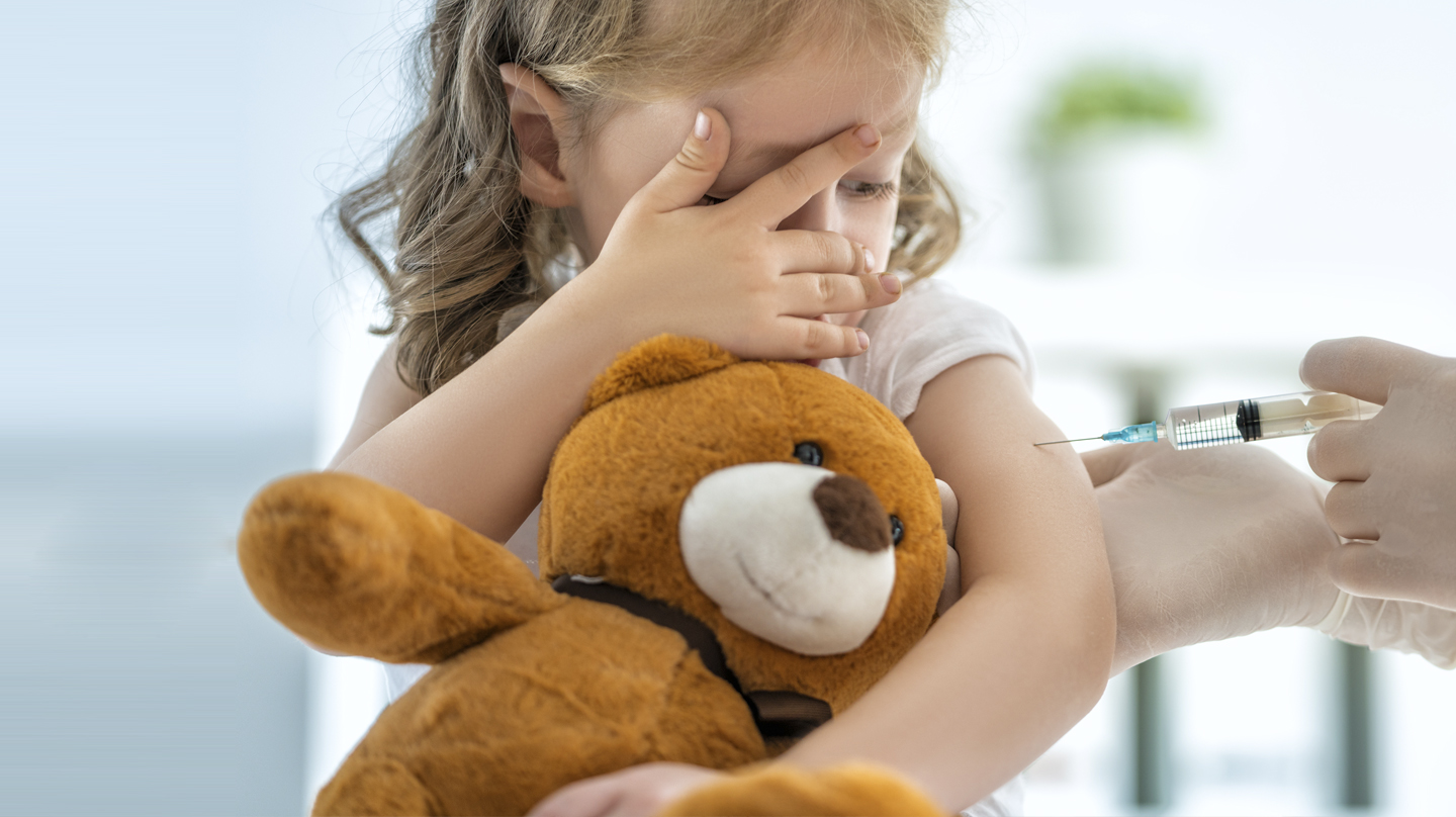 Angst vor Spritzen: Kleines Mädchen mit Teddybär im Arm schaut skeptisch auf die Spritze, die ihr der Arzt gerade verabreicht.